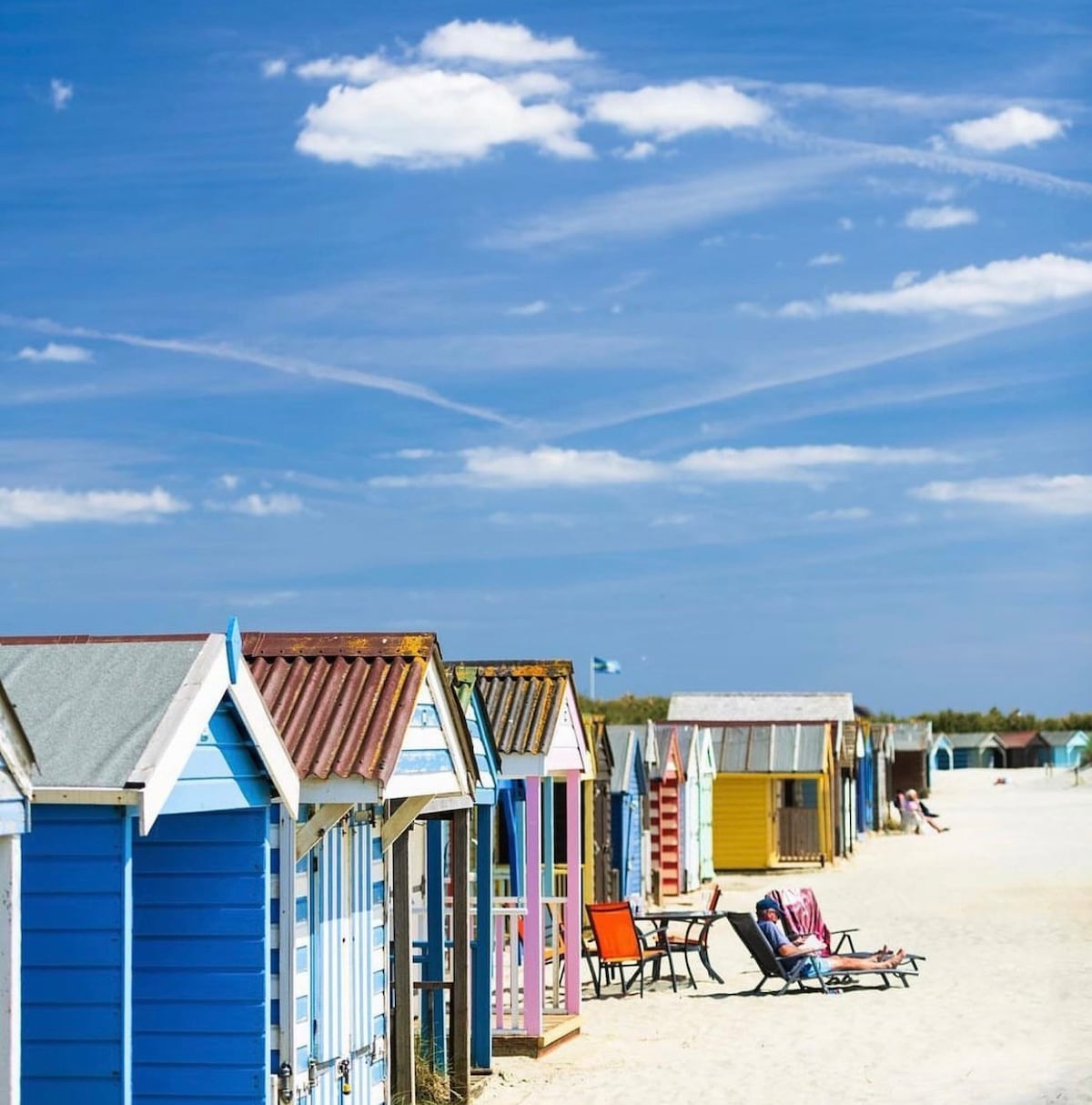 A row of colorful beach huts is positioned along a sandy beach, with various shades of blue, yellow, and pink visible. In the foreground, a seated individual enjoys the sun, surrounded by warm sand and clear blue skies dotted with fluffy white clouds.