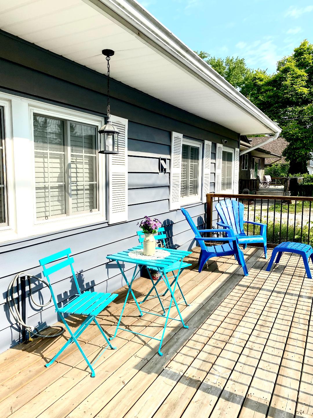 A deck area features a small blue bistro table accompanied by matching chairs, creating a relaxed outdoor seating space. Bright sunlight illuminates the grey siding of the cottage, and a lantern hangs nearby, enhancing the inviting environment.