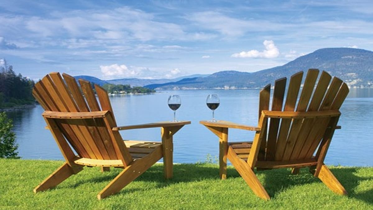 Two wooden Adirondack chairs are positioned on a green lawn, facing Okanagan Lake. Each chair has a glass of wine placed on a small table between them, with a scenic view of the calm lake and rolling hills in the background.
