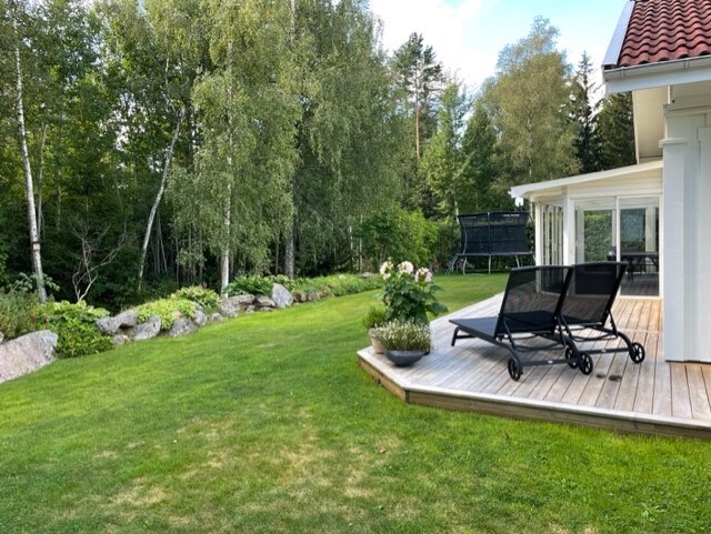 A spacious outdoor area features a wooden deck lined with lounge chairs, surrounded by a lush green lawn. Trees and shrubs create a natural backdrop, with decorative plants placed near the house. An adjacent play area with a trampoline can be seen in the distance.
