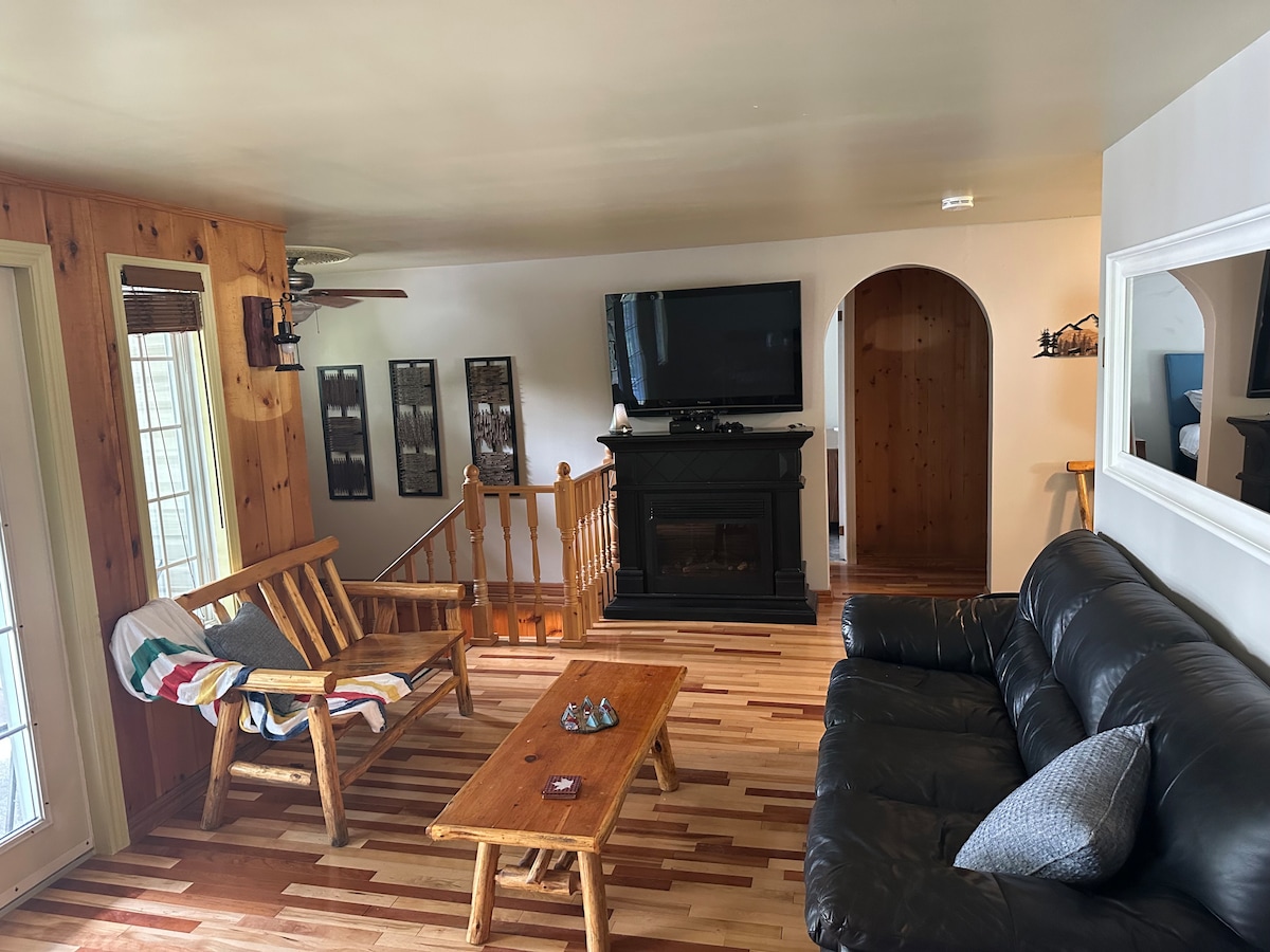 A welcoming living area features a mix of wood and modern design. A dark sofa and rustic-style chairs are arranged around a wooden coffee table. A large television is mounted above a dark fireplace, and natural light pours in from large windows.