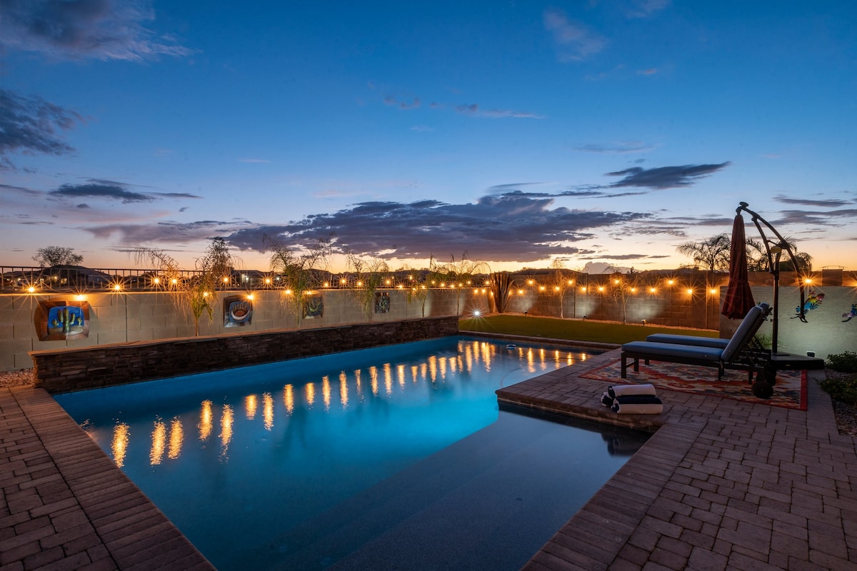 A serene outdoor pool area is shown, with a crystal-clear pool reflecting evening lights. The space is bordered by a decorative wall, with gentle landscaping and cozy seating areas. String lights illuminate the surroundings, enhancing the atmosphere for relaxation.