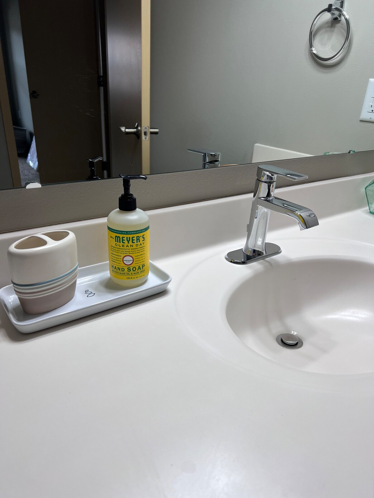 A bathroom countertop is depicted, featuring a sleek faucet beside a white sink. A yellow hand soap dispenser and a small cotton holder are positioned on a tray. A mirror reflects the spacious area, while a doorway leads to another room in the background.