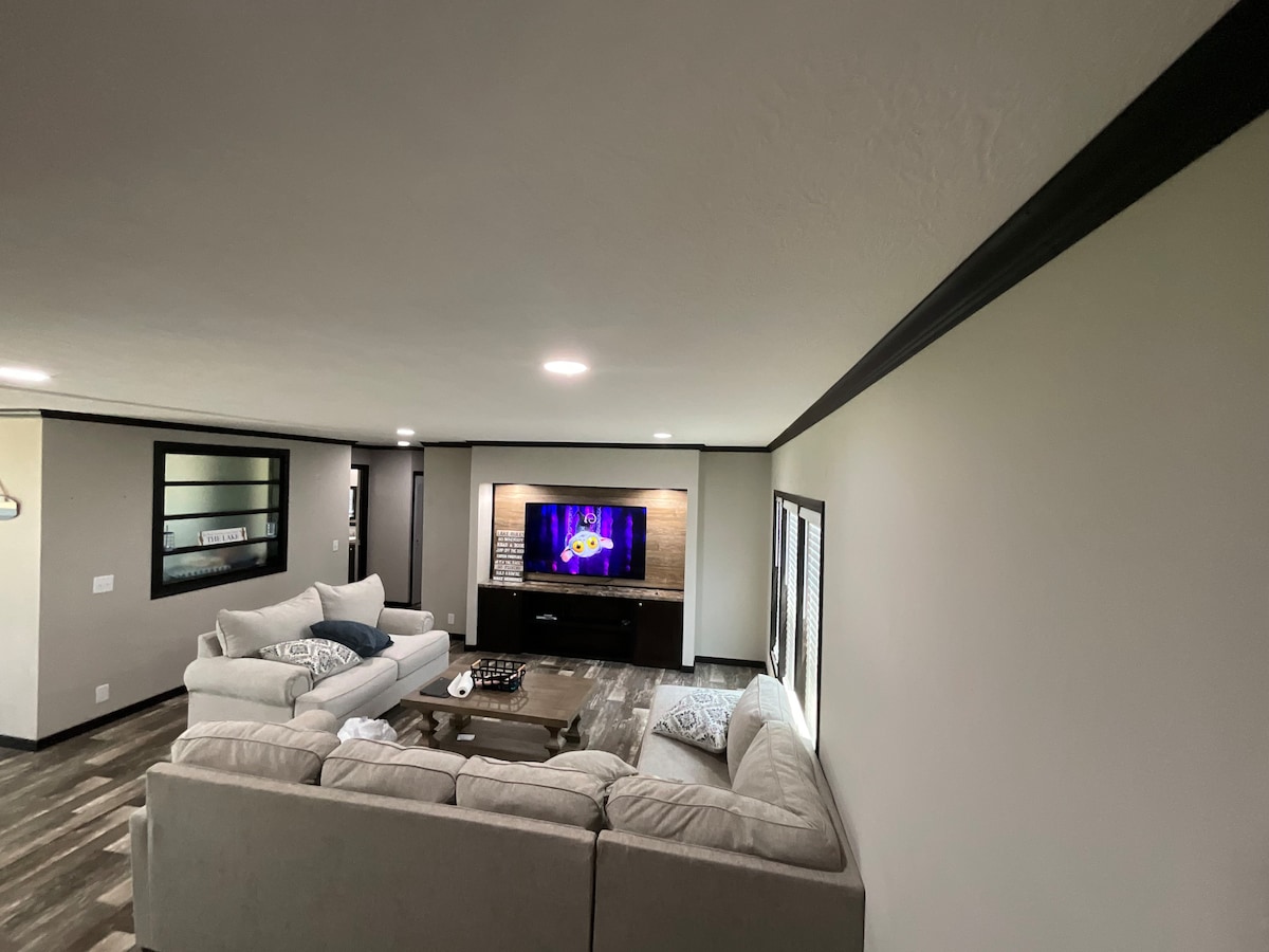 A spacious living area features a large sectional sofa and a cozy armchair. A wooden coffee table is centered on a textured rug. A wall-mounted TV is displayed above a modern media console, with soft lighting creating an open and inviting atmosphere.