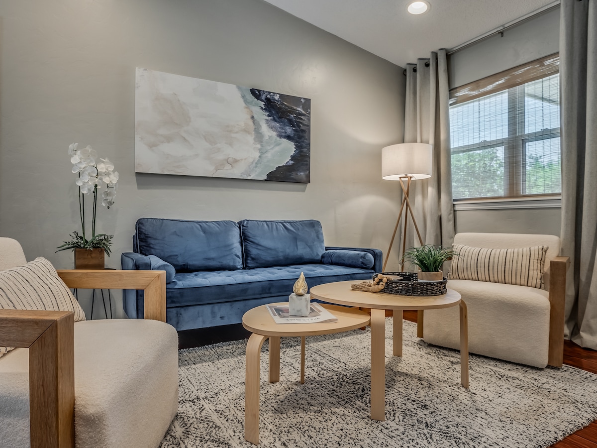 A comfortable seating area is featured, including a blue sofa and two light-colored chairs. A coffee table with decorative items sits at the center, surrounded by a textured rug. Natural light enters through a window adorned with light curtains, illuminating the calming space.