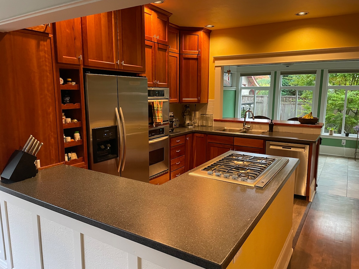 A spacious kitchen features wooden cabinetry illuminated by warm lighting. A stainless steel refrigerator and oven are prominently positioned, while a gas cooktop is integrated into the counter. The kitchen offers a view of the garden through large windows, enhancing the inviting atmosphere.