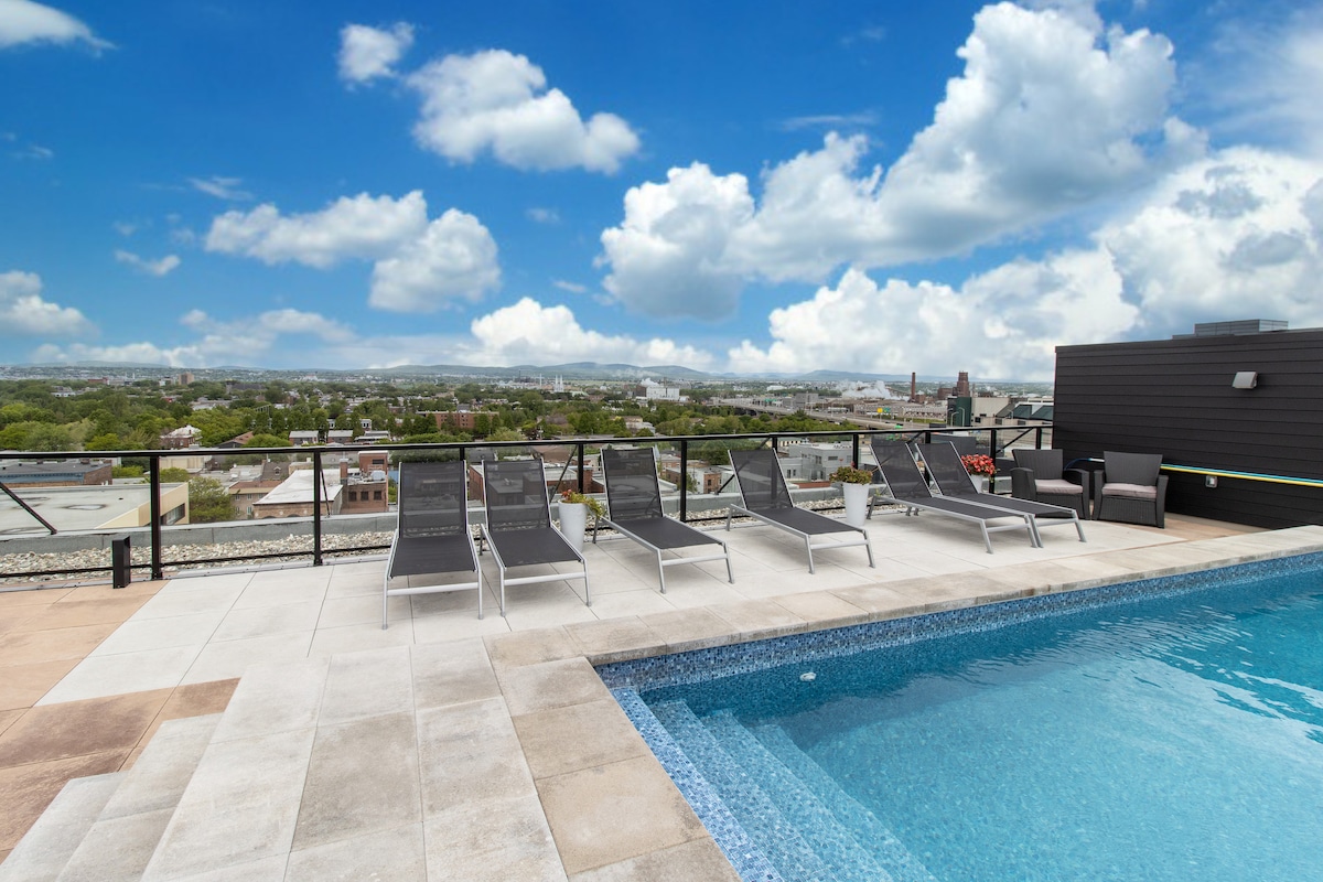 A rooftop area features a swimming pool surrounded by multiple lounge chairs. The glass railing provides an uninterrupted view of the surrounding cityscape and sky, creating a serene atmosphere for relaxation.