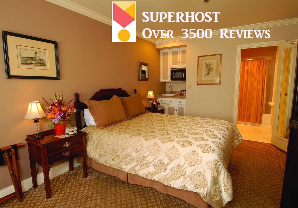 A cozy hotel room features a spacious bed adorned with a decorative cover. A small kitchenette is visible, offering convenient amenities. Soft lighting from bedside lamps adds warmth, while a private bathroom is accessed through a door in the background. Floral decor adds a touch of color.