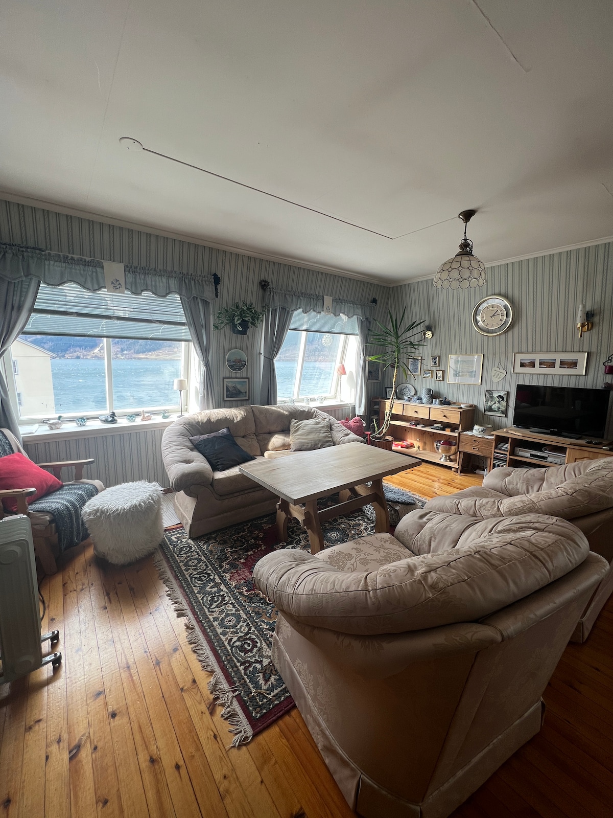 A spacious living area features comfortable seating arranged around a wooden coffee table. Large windows showcase scenic views of the fjord, while natural light illuminates the room. Decorative elements include plants and wall art, adding character to the cozy atmosphere.