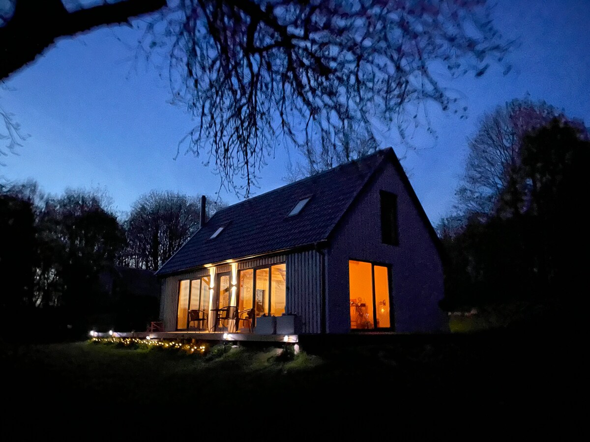 The house features large glass windows, revealing a warm glow from within. The exterior is surrounded by trees, with a well-maintained lawn and subtle outdoor lighting that guides the path. A tranquil evening atmosphere is suggested by the darkening sky.