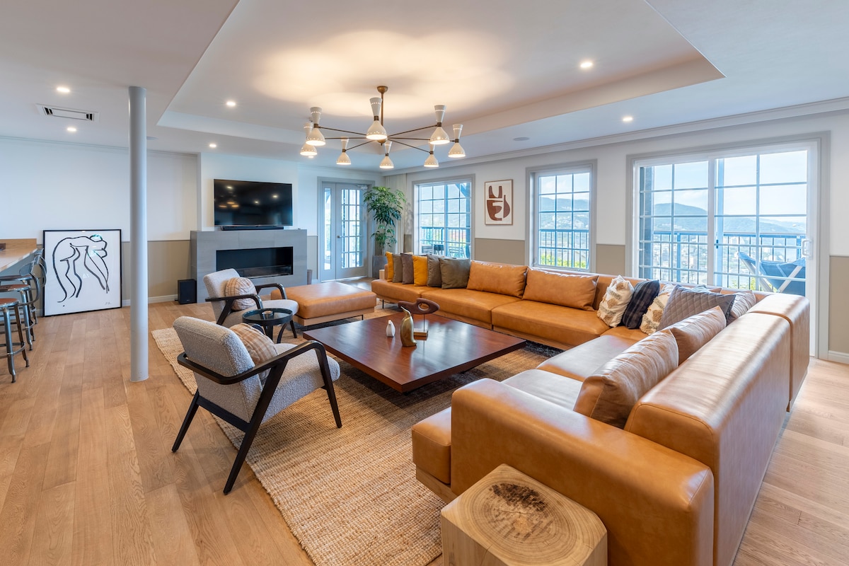 A spacious living area is designed with natural light flooding through large windows, featuring a large sectional sofa surrounding a central wooden coffee table. A flat-screen TV is mounted on the wall, complemented by a subtle artwork to the side.