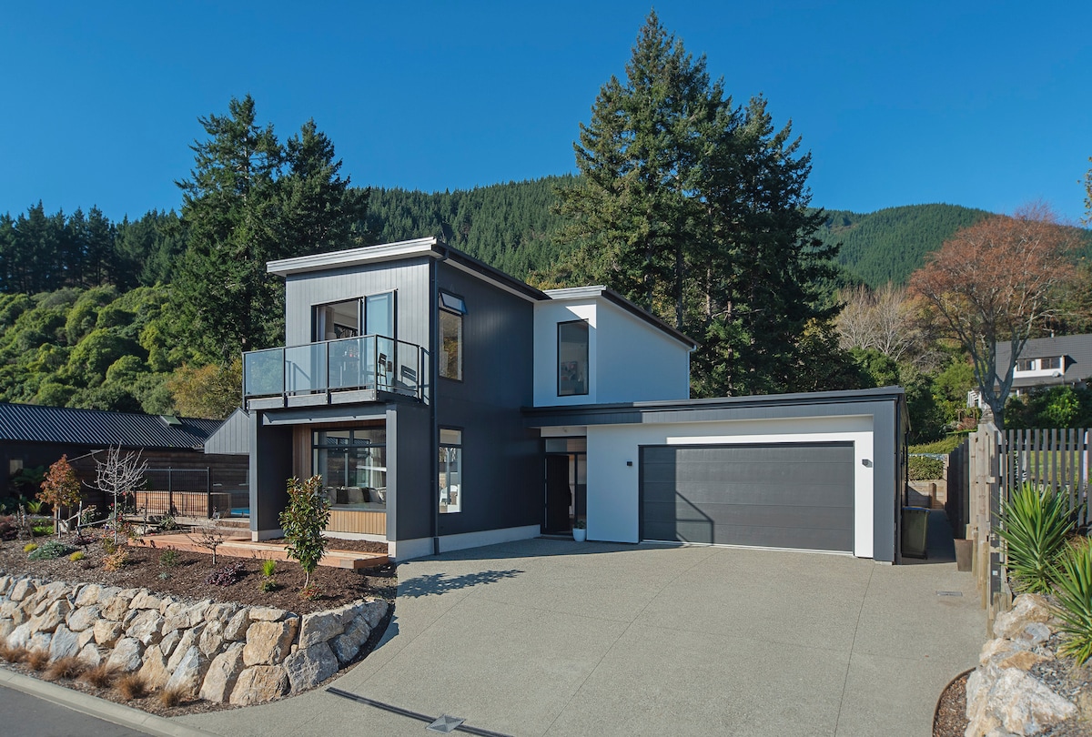 A newly built holiday home is presented with a modern exterior and large front windows. The two-story design features a balcony on the upper level, surrounded by trees and greenery, while a well-maintained driveway leads to a closed garage.