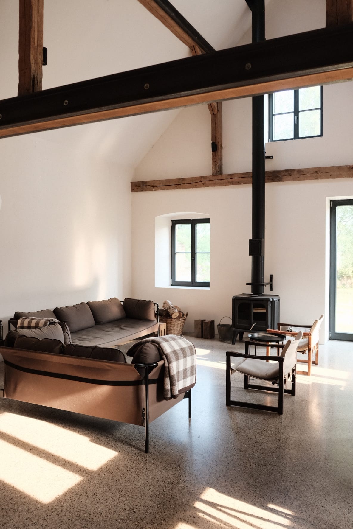 A spacious living area features a comfortable sectional sofa adorned with a lightweight throw. Natural light filters through large windows, highlighting the polished concrete floor. A compact wood stove is positioned nearby, complemented by simple wooden chairs and a cozy basket, enhancing a relaxed atmosphere.