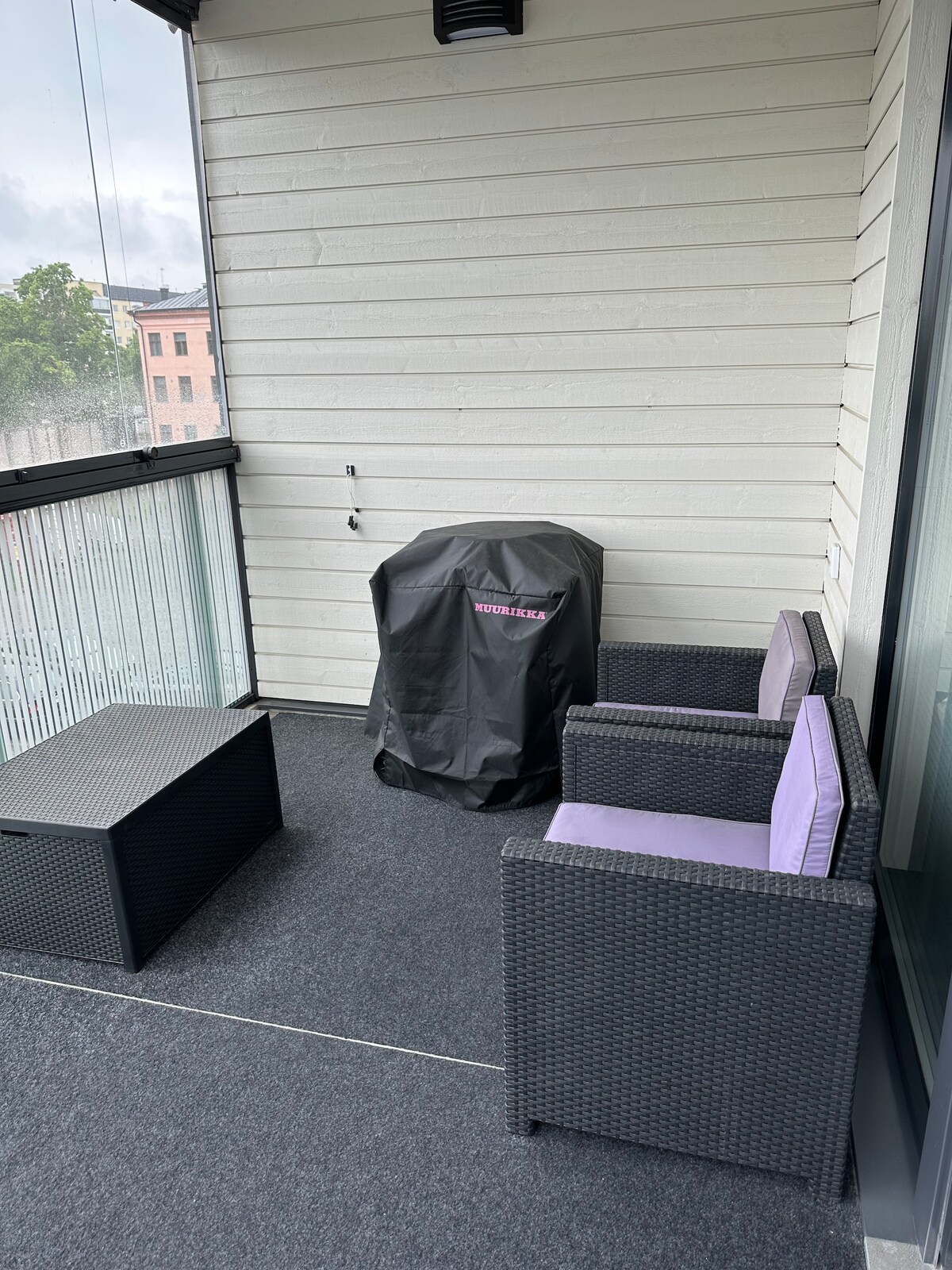 A balcony area is seen featuring two wicker chairs with purple cushions and a low table. A covered grill sits in one corner, surrounded by glass panels that provide a view of the surroundings.