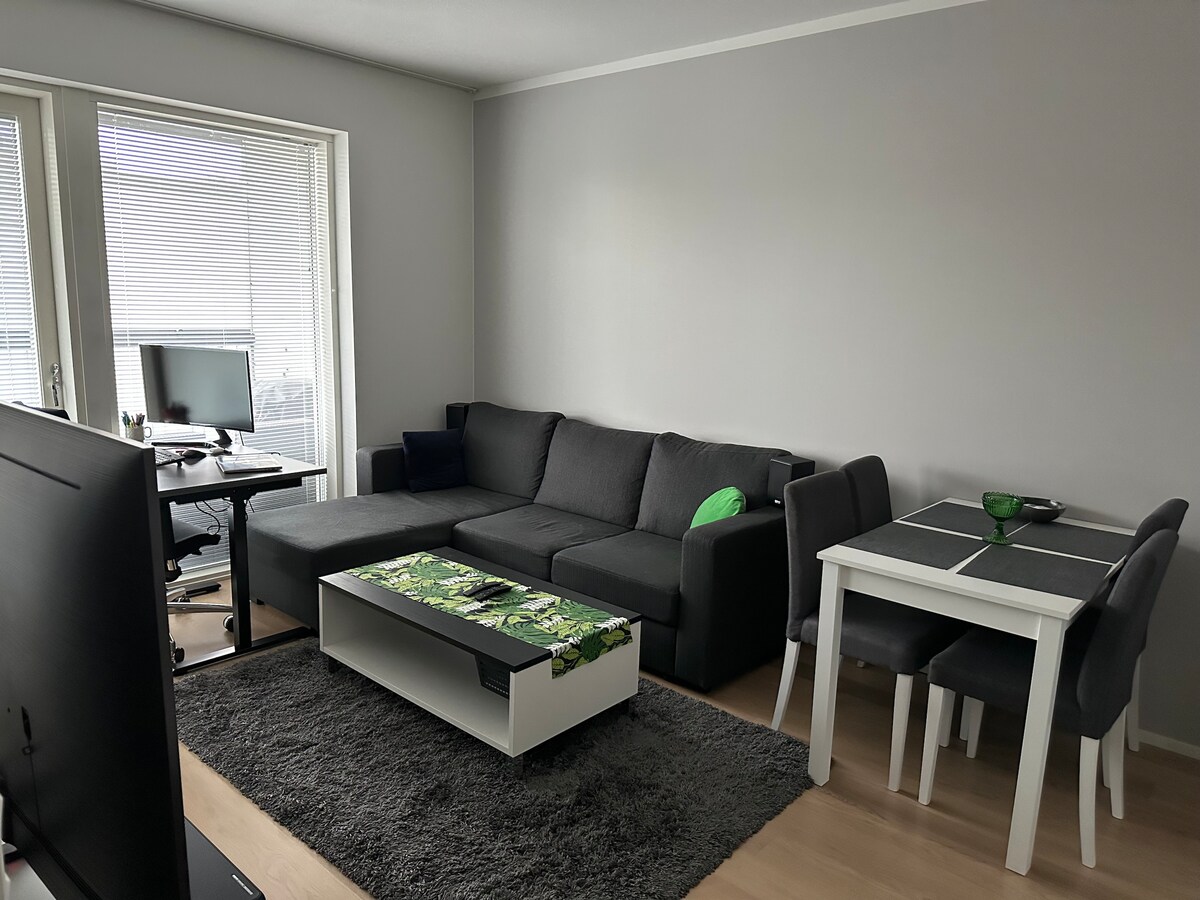 A modern living space is presented with a dark sectional sofa and a coffee table featuring a decorative top. A dining table with four chairs is positioned nearby. Natural light enters through large windows, enhancing the neutral color palette of the room.