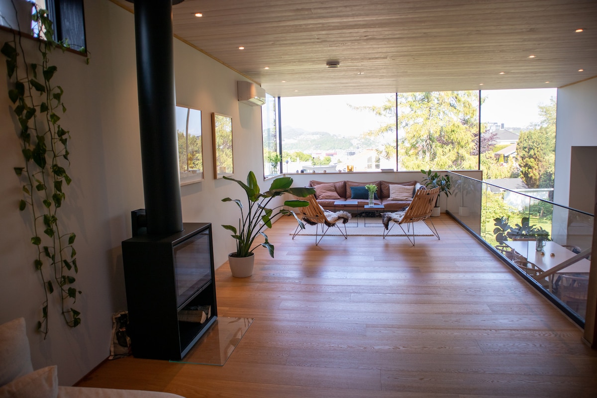 A bright living area is defined by large glass windows that offer views of the surrounding greenery. A spacious seating arrangement is complemented by natural wooden flooring and a modern fireplace. Decorative plants add a touch of nature to the inviting environment.