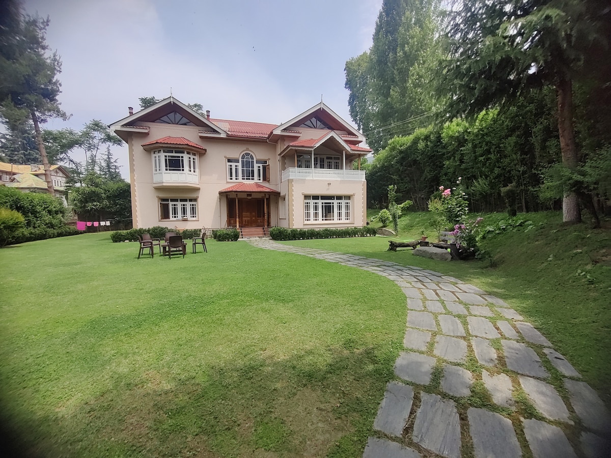 The exterior of a spacious villa is shown, featuring a well-maintained lawn surrounded by trees. A stone pathway leads to the entrance, while outdoor seating is arranged nearby. The house has a traditional design with large windows and a charming red-roof, blending harmoniously with the landscape.