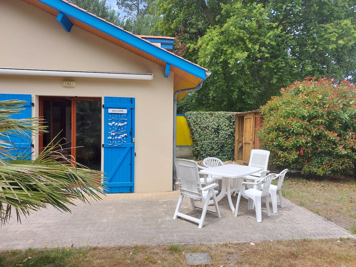 An outdoor terrace is shown, featuring a dining table surrounded by white chairs. Bright blue shutters adorn the entrance, while greenery offers a natural backdrop. A shaded area from a retractable awning is visible, contributing to a relaxed outdoor setting.