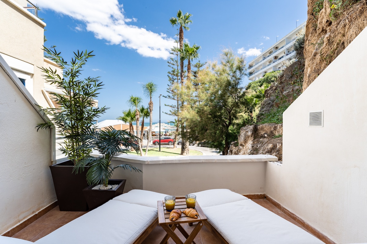 A spacious terrace is featured with two comfortable sun loungers positioned to enjoy the view. Lush greenery is present, along with a small table set with two glasses and pastries. The backdrop includes palm trees and coastal scenery under a clear blue sky.