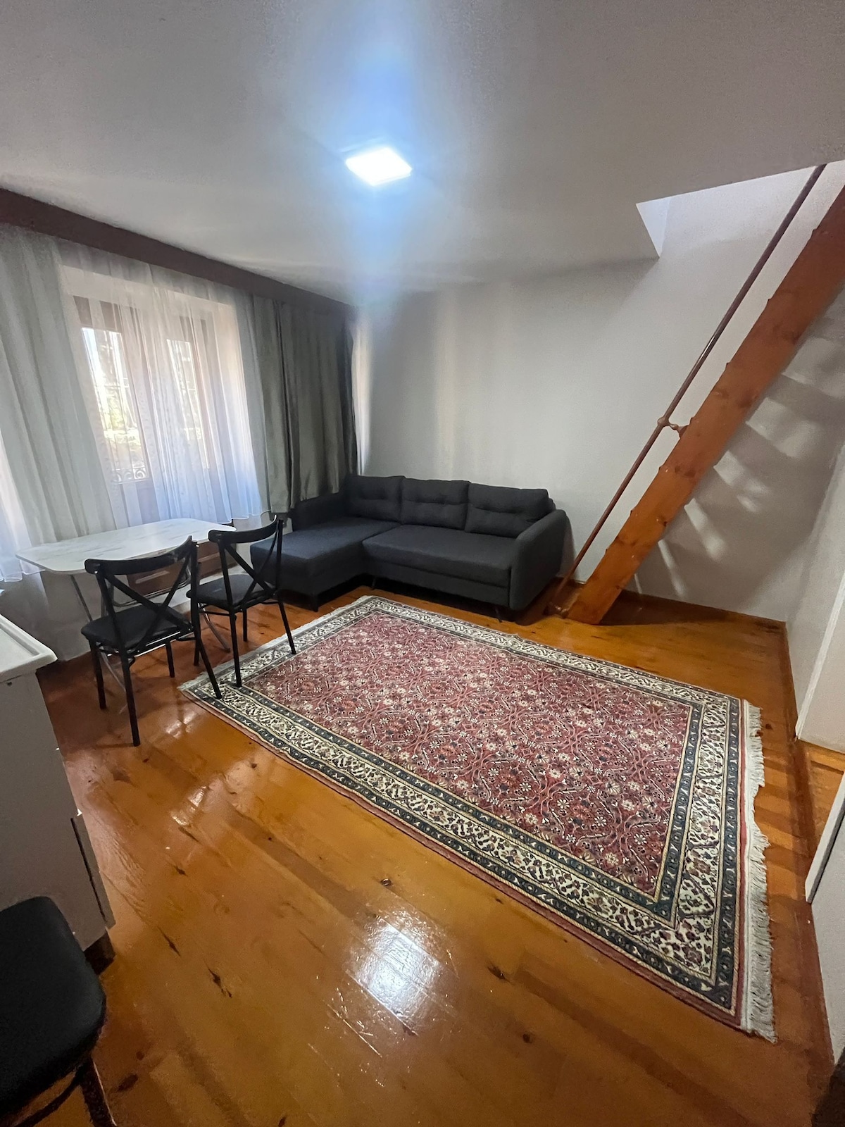 A cozy living area features a dark sectional sofa, complemented by a wooden staircase. A small dining table with black chairs is situated near a window, allowing natural light to fill the space. A decorative area rug adds warmth to the wooden floor.