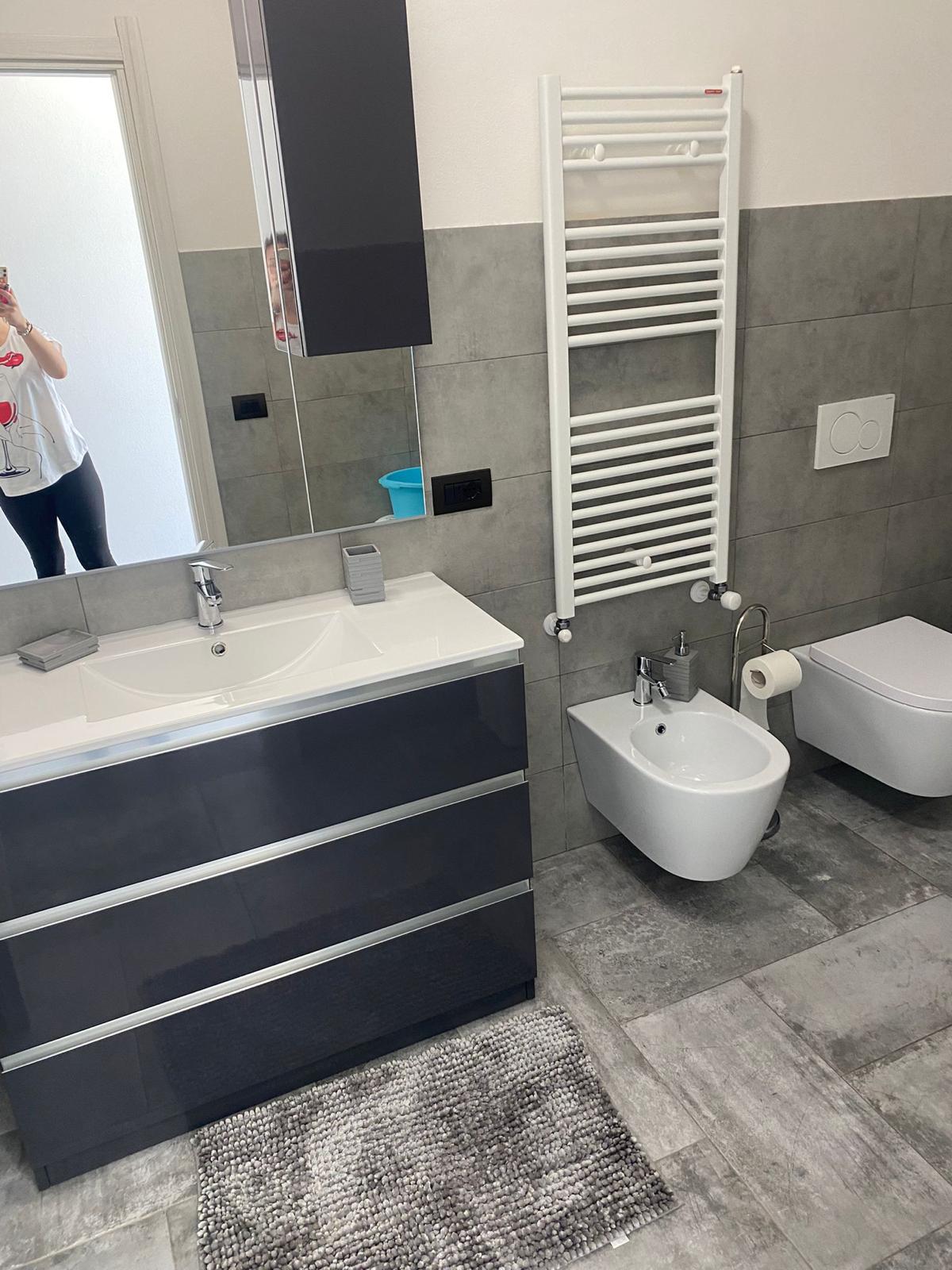The bathroom features a modern design with gray tiles. A sleek vanity with multiple drawers and a mirror above is complemented by a bidet and a wall-mounted toilet. A heated towel rack is positioned beside the fixtures, and a textured rug is placed on the floor.