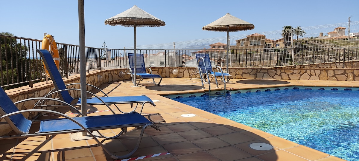A sunlit pool area is highlighted by a refreshing blue pool surrounded by a tiled deck. Four lounge chairs offer comfort under shaded umbrellas, inviting relaxation. The surrounding landscape features residential structures and gentle hills, creating a calm and inviting environment.
