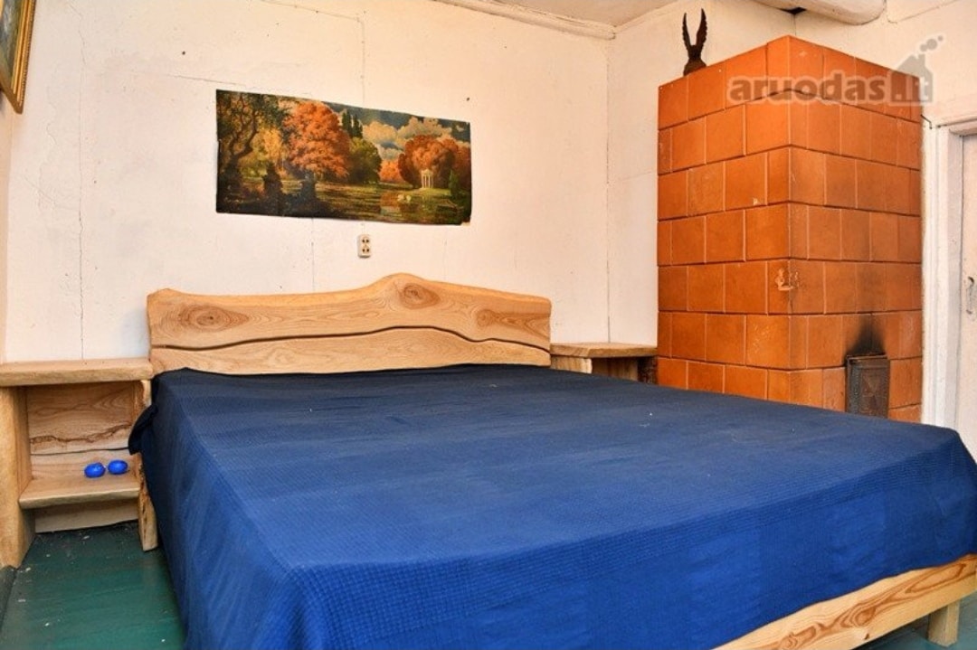 A spacious bed is positioned against a wooden wall, featuring a deep blue blanket. A painting depicting a serene landscape hangs above the bed. Nearby, a warm-colored tiled stove adds a rustic touch to the room.