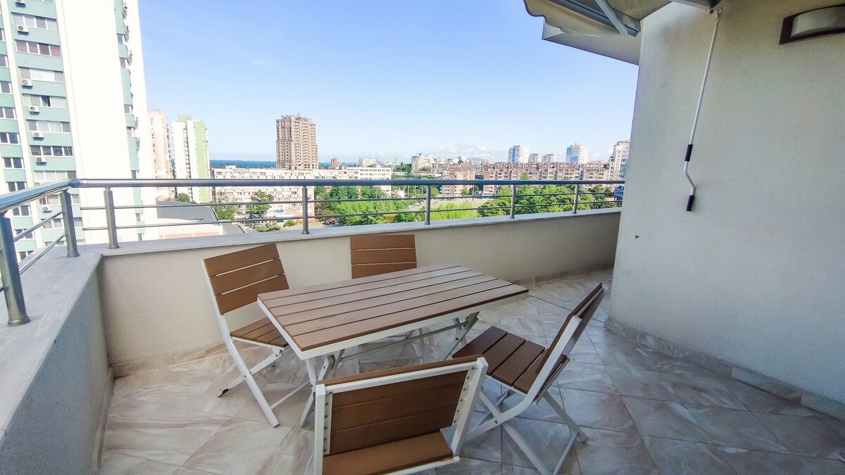 An outdoor terrace features a table surrounded by four chairs, offering panoramic views of the cityscape and horizon. The space is designed for al fresco dining and relaxation, with sunlight illuminating the tiled floor.