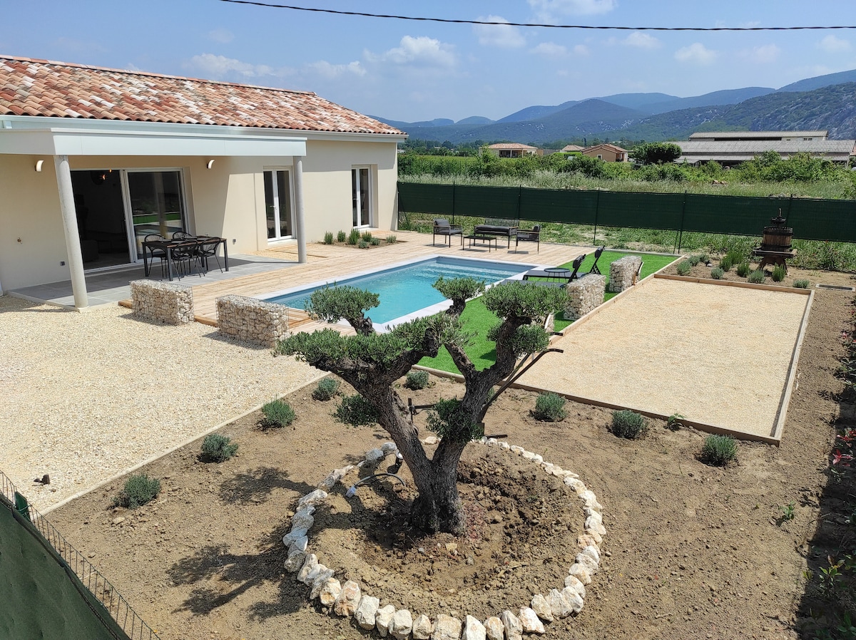An outdoor area features a private swimming pool surrounded by landscaped grounds. A stone path leads to a central olive tree with low stone borders. Comfortable seating is arranged near the pool, with open views of the surrounding hills under a clear sky.