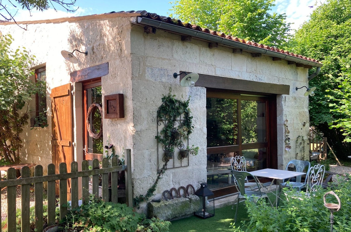 A charming stone house is depicted, showcasing large glass doors framed by wooden accents. A small outdoor dining area with a table and chairs is visible, surrounded by lush greenery and garden elements. Warm sunlight highlights the inviting façade.