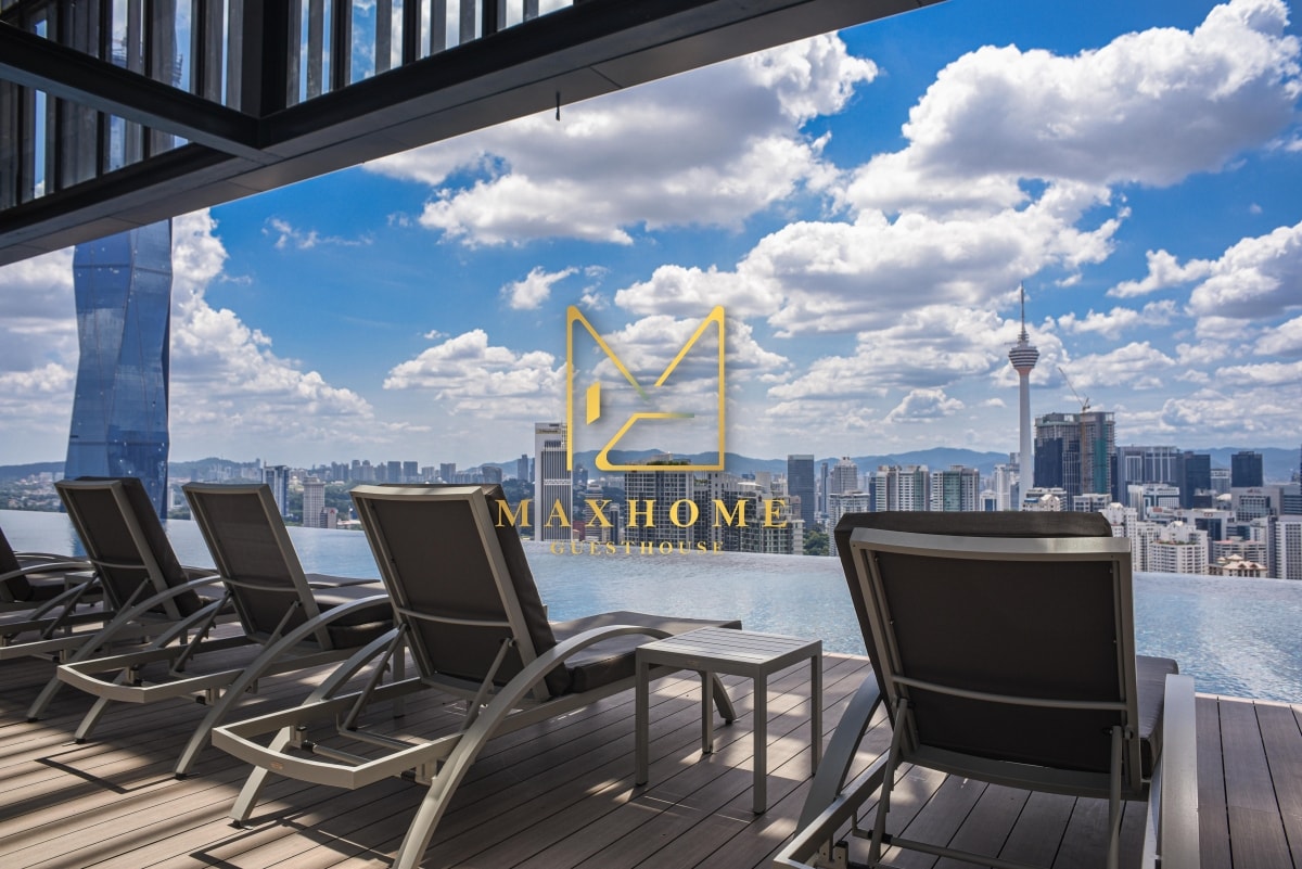 Lounge chairs are positioned on a wooden deck, overlooking a panoramic city skyline. The scene features a refreshing swimming pool with stunning views of tall buildings and the iconic KL Tower under a partly cloudy sky.