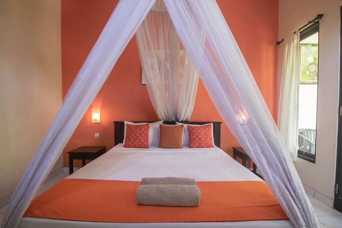 A spacious bed is draped in white fabric, creating an airy feel. The room features a warm orange accent wall, complemented by soft lighting. Two side tables are positioned beside the bed, while a neatly rolled towel rests at the foot. Natural light filters through the nearby window.
