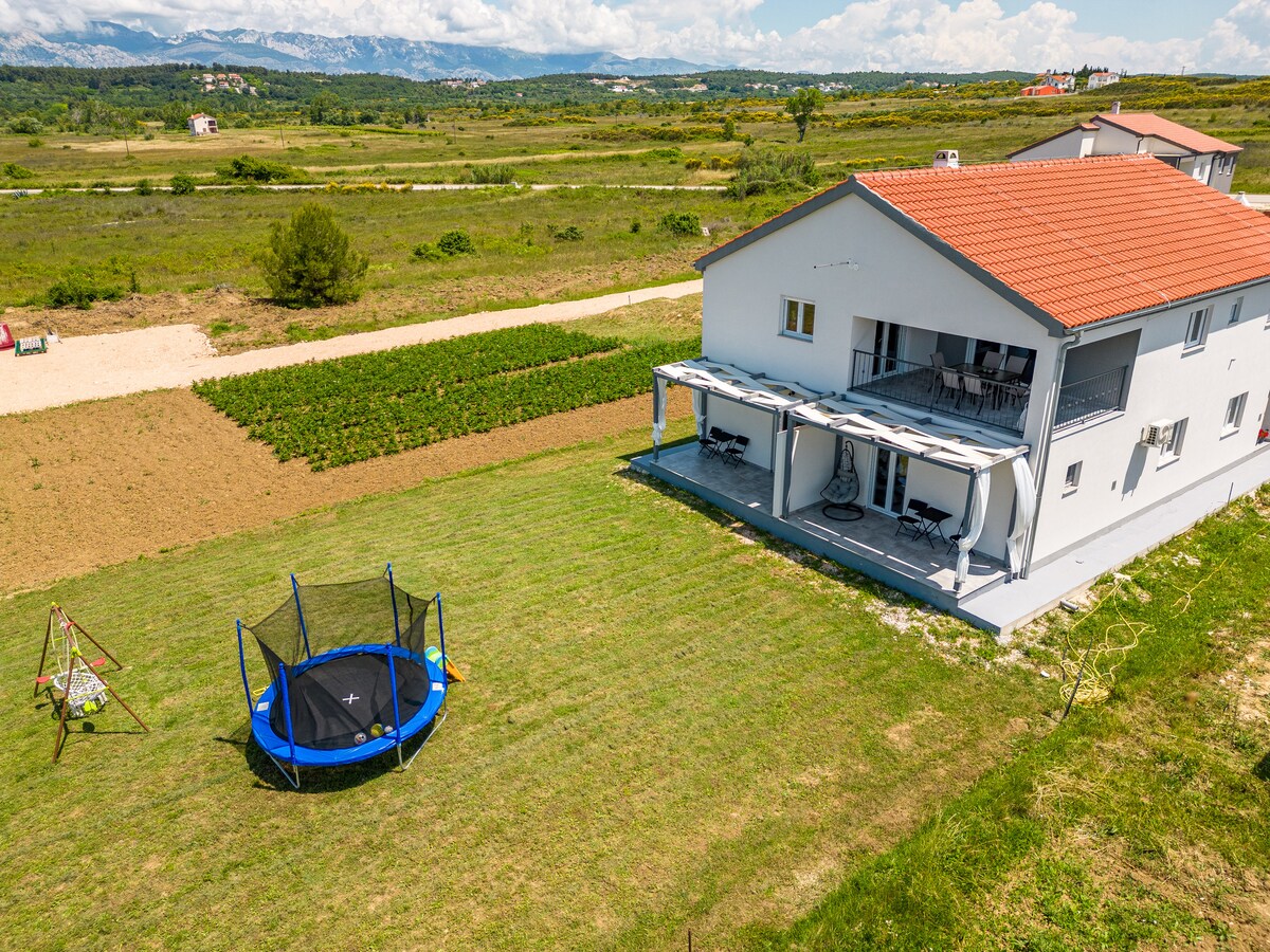 An outdoor view reveals a spacious green lawn featuring a trampoline and a swing set. In the background, a modern two-story structure is complemented by a covered patio. A scenic landscape with distant hills and a clear blue sky can be observed.