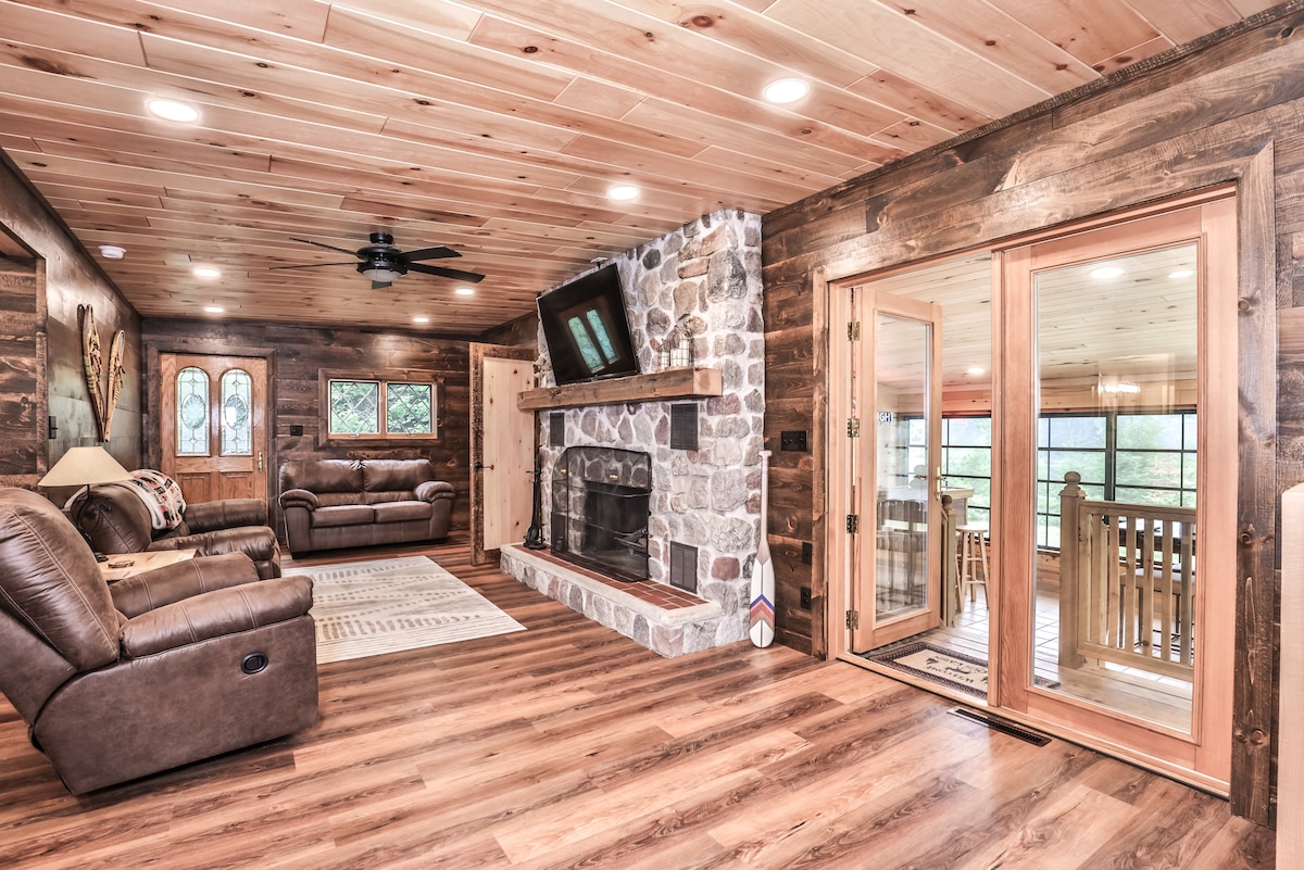 A spacious living area features warm wooden paneling, comfortable leather couches, and a stone fireplace as a centerpiece. Large windows invite natural light. A flat-screen TV is mounted above the fireplace, and doors lead to an outdoor space, enhancing indoor-outdoor flow.