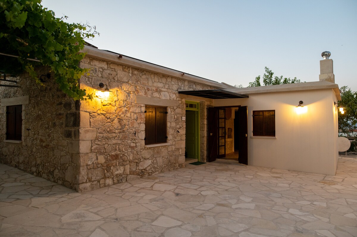 The exterior of the bungalow features traditional stone architecture with a warm glow from outdoor lighting. The entrance is flanked by wooden windows, and a spacious stone patio is visible, surrounded by greenery.