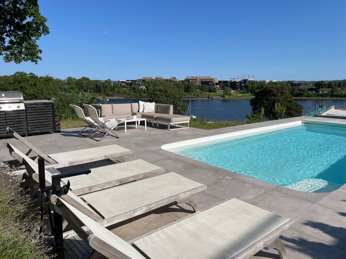 A modern outdoor space is presented, featuring a private swimming pool surrounded by a spacious deck. Comfortable seating arrangements are included, along with sun loungers. A scenic view of the nearby water and greenery enhances the backdrop.