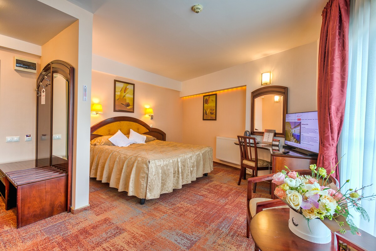 A spacious hotel room features a large bed adorned with a golden bedspread. Soft lighting from wall sconces creates a serene ambiance. A desk is positioned near a window, alongside a comfortable chair and a small table holding a vase of fresh flowers.