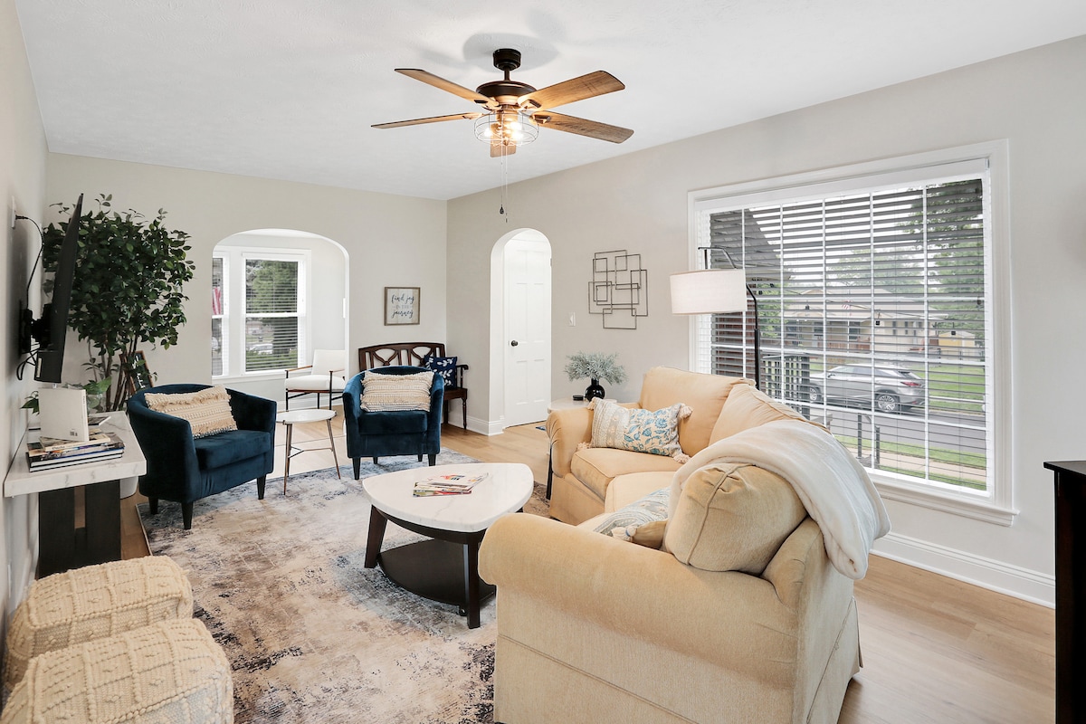 A comfortable living room is presented, featuring a spacious sofa and two upholstered chairs. Natural light filters through large windows adorned with blinds. A decorative area rug covers the hardwood floor, while a ceiling fan adds to the inviting ambiance.