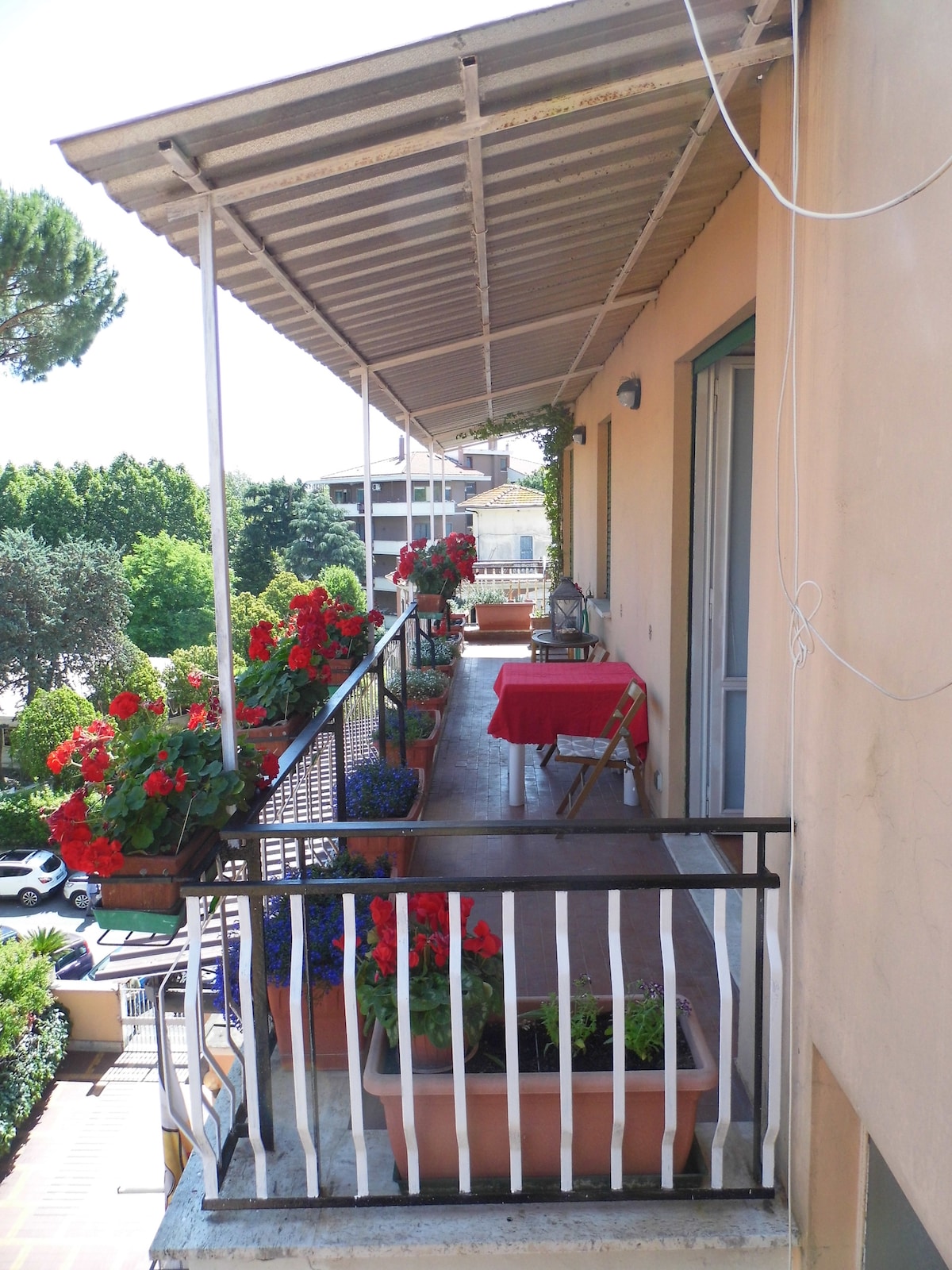 A balcony adorned with vibrant red flowers in planters creates a lively atmosphere. A small table covered with a red cloth is positioned at one end, providing a cozy spot to enjoy the outdoors. Green trees and buildings are visible in the background.