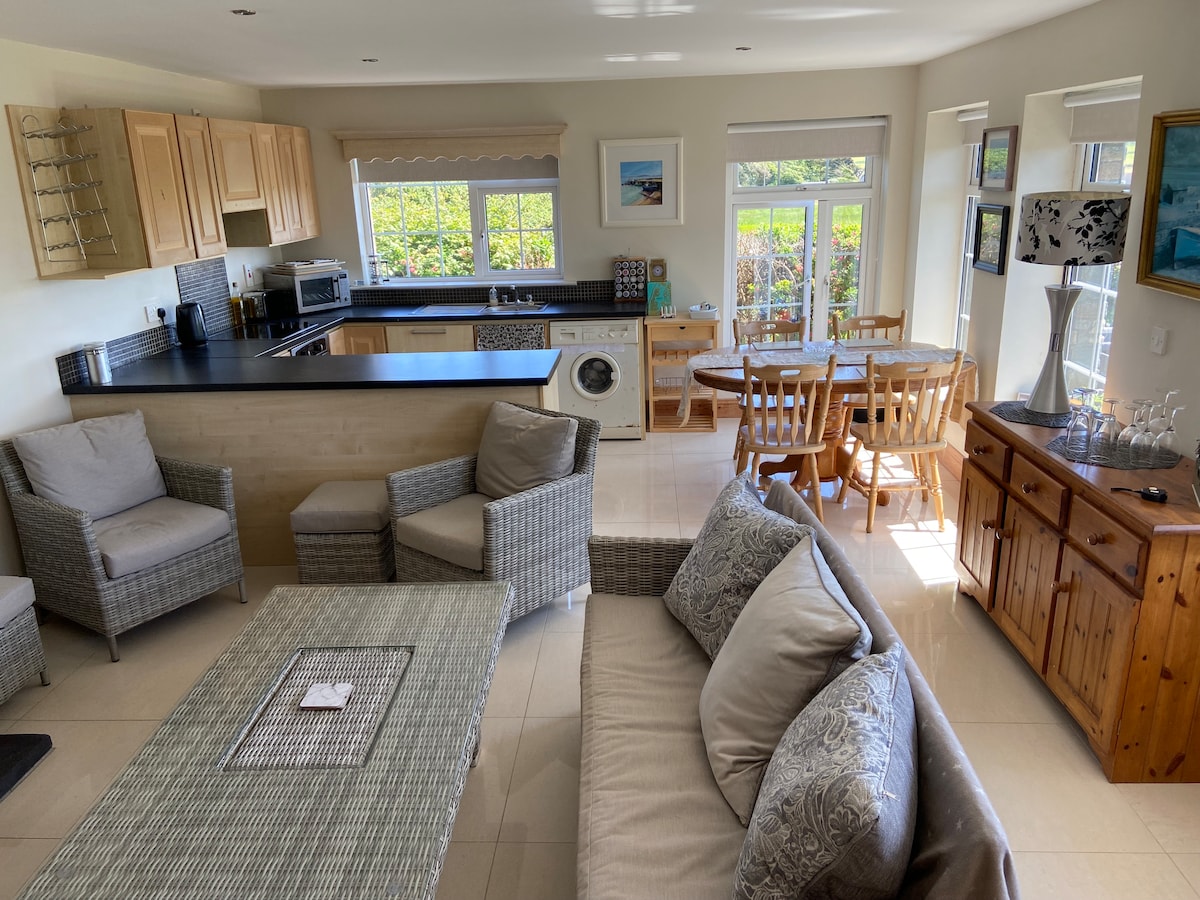 The open-plan living area features wicker seating and a spacious coffee table. A well-equipped kitchen is visible in the background, complemented by a dining table with seating for six. Natural light fills the room through large windows overlooking a garden.