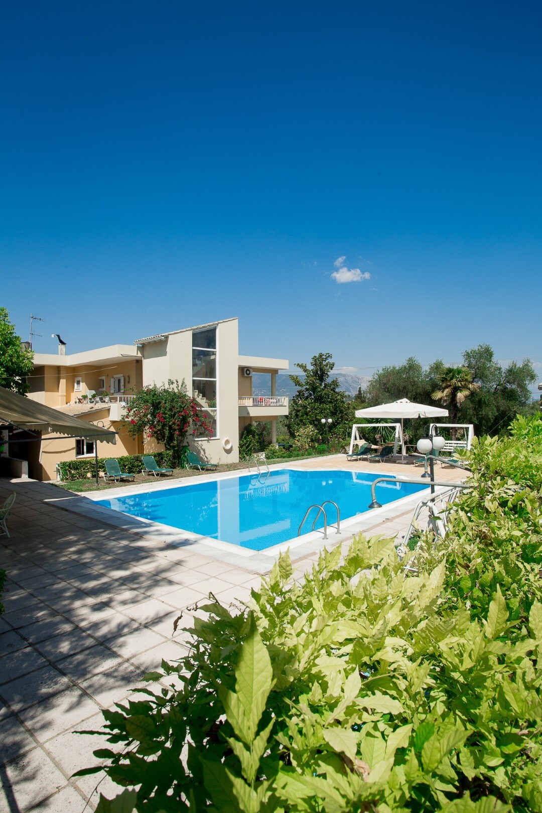 A refreshing swimming pool, surrounded by a well-maintained garden, is situated in front of a two-story building. Sun loungers are placed on the poolside deck, offering leisure space. Lush greenery and flowering plants add a natural touch to the serene environment under a clear blue sky.