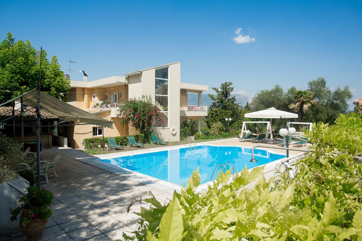 A spacious outdoor area features a rectangular swimming pool surrounded by lounge chairs. A private gazebo is positioned nearby, with greenery visible throughout the garden. The two-story building stands in the background, complemented by blooming flowers and sprawling trees under a clear blue sky.