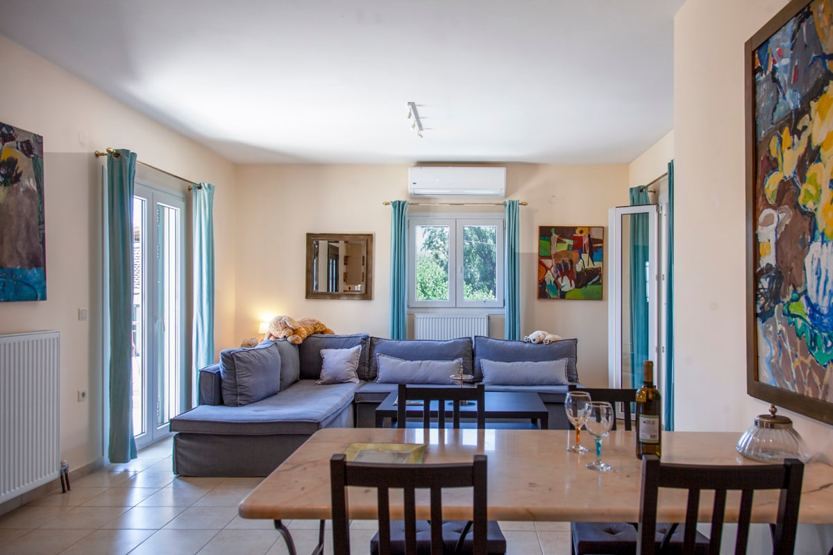 A spacious living area is presented with a sectional sofa, coffee table, and dining table for four. Light enters through large windows, highlighting colorful artwork on the walls. A sliding door leads to outdoor space, enhancing the comfortable atmosphere.