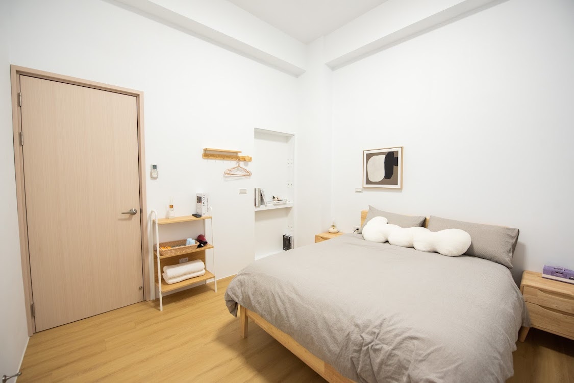 A cozy room features a spacious bed with light bedding against a neutral wall. A wooden door leads outside, while a small shelf and a hanging rack are visible. A minimalist artwork adds a touch of design, complementing the overall serene ambiance.