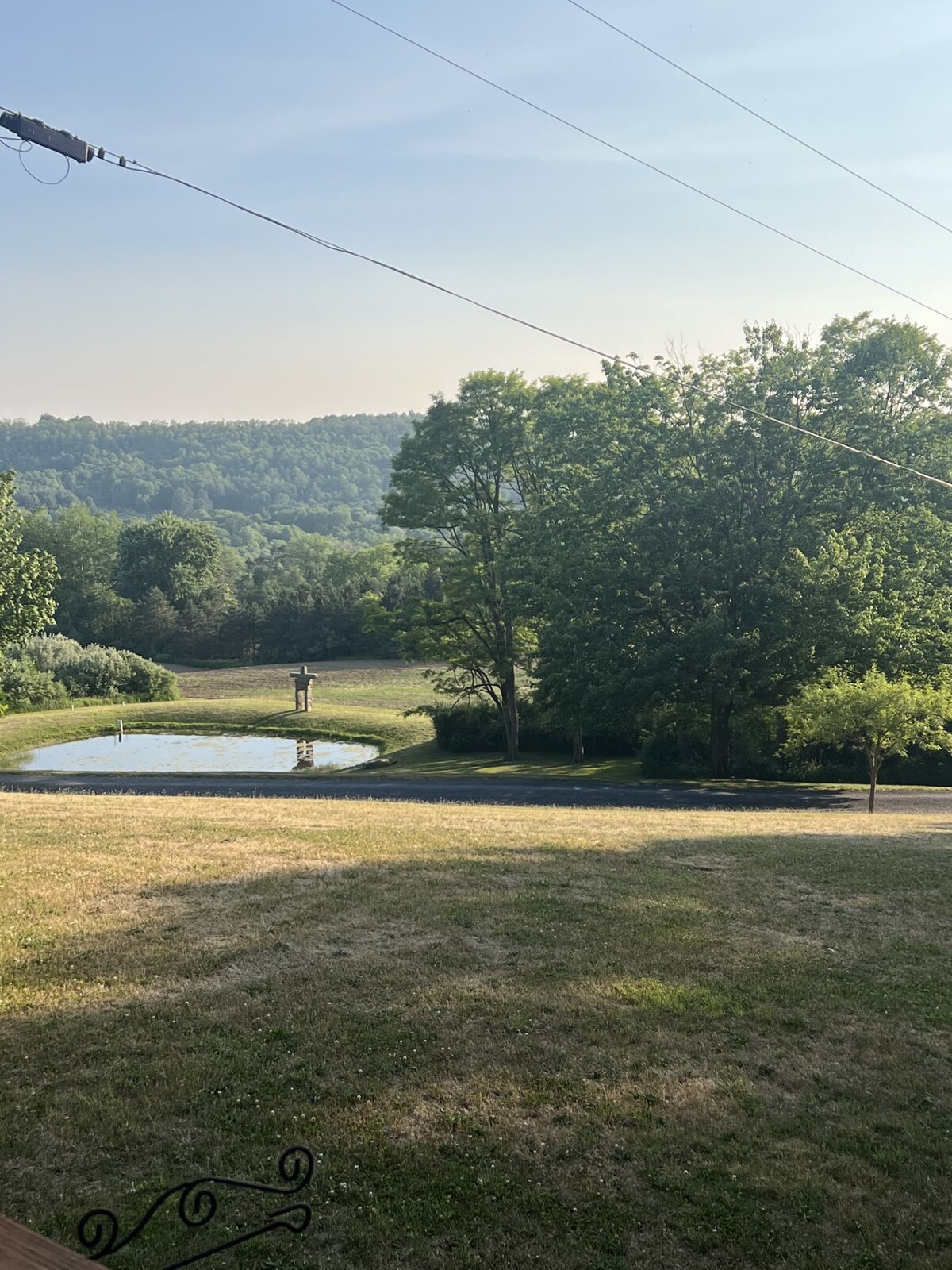A serene landscape reveals a clear pond surrounded by grassy areas and mature trees. Rolling hills can be seen in the background, offering a natural setting that embodies tranquility. A pathway leads from the house toward the scenic view, enhancing the outdoor experience.