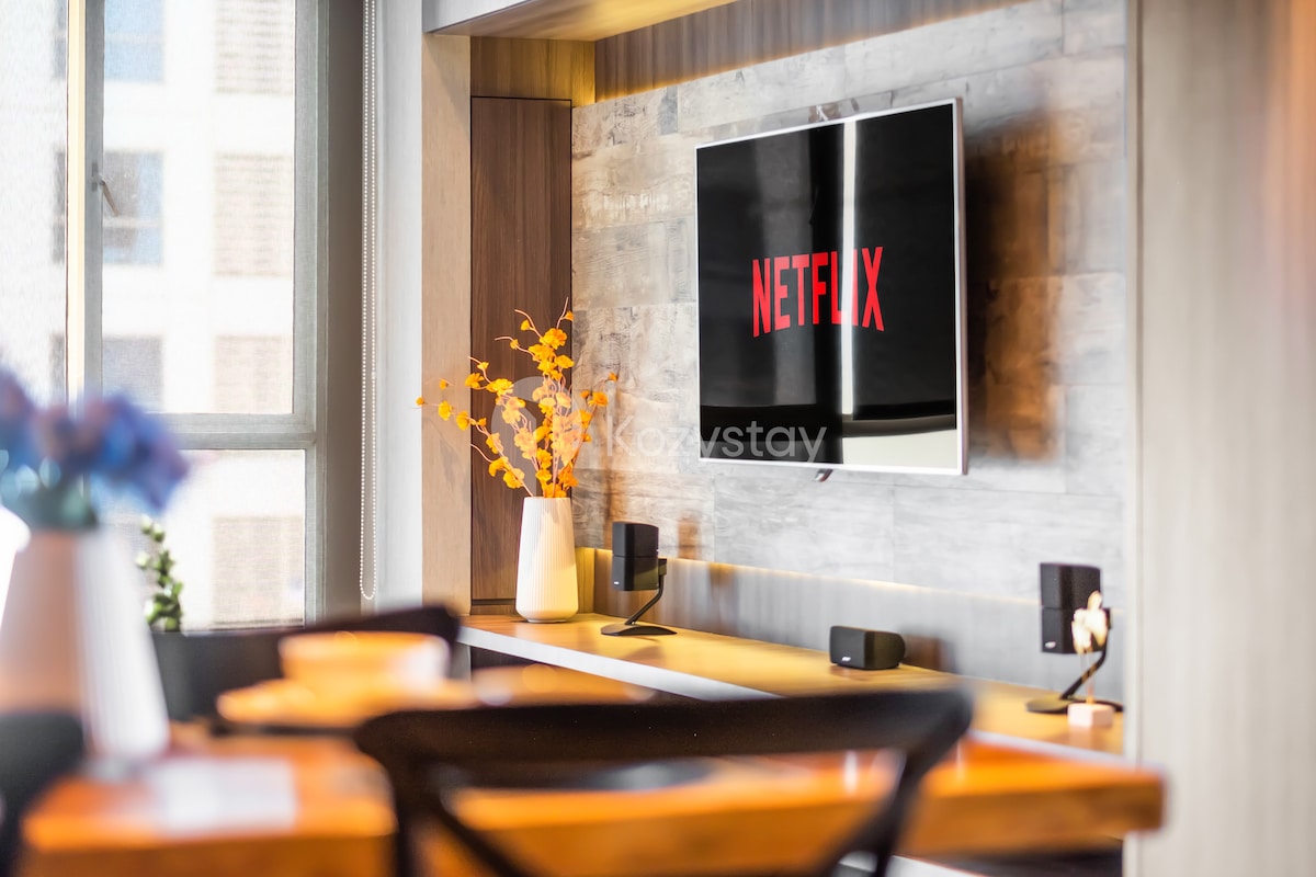 A modern living space features a wall-mounted television displaying the Netflix logo. A wooden shelf below holds a speaker and decorative vase with orange flowers. Soft light illuminates the area, with a dining table visible in the foreground.