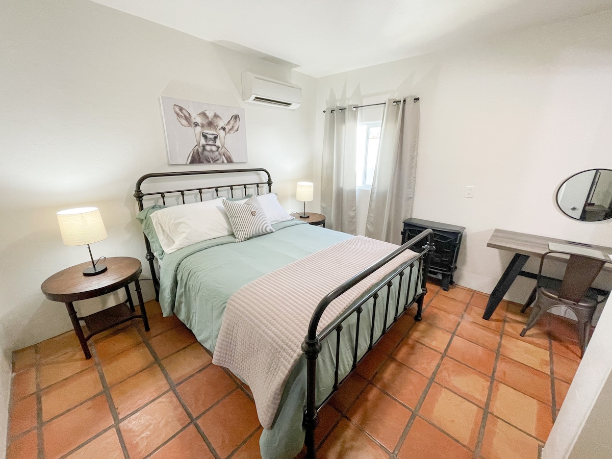 A cozy bedroom features a queen bed with a light blue blanket and decorative throw. Two bedside lamps provide soft lighting. A window, adorned with light curtains, and a mirror beside a small desk enhance the room's functionality. The floor is tiled in warm terracotta.