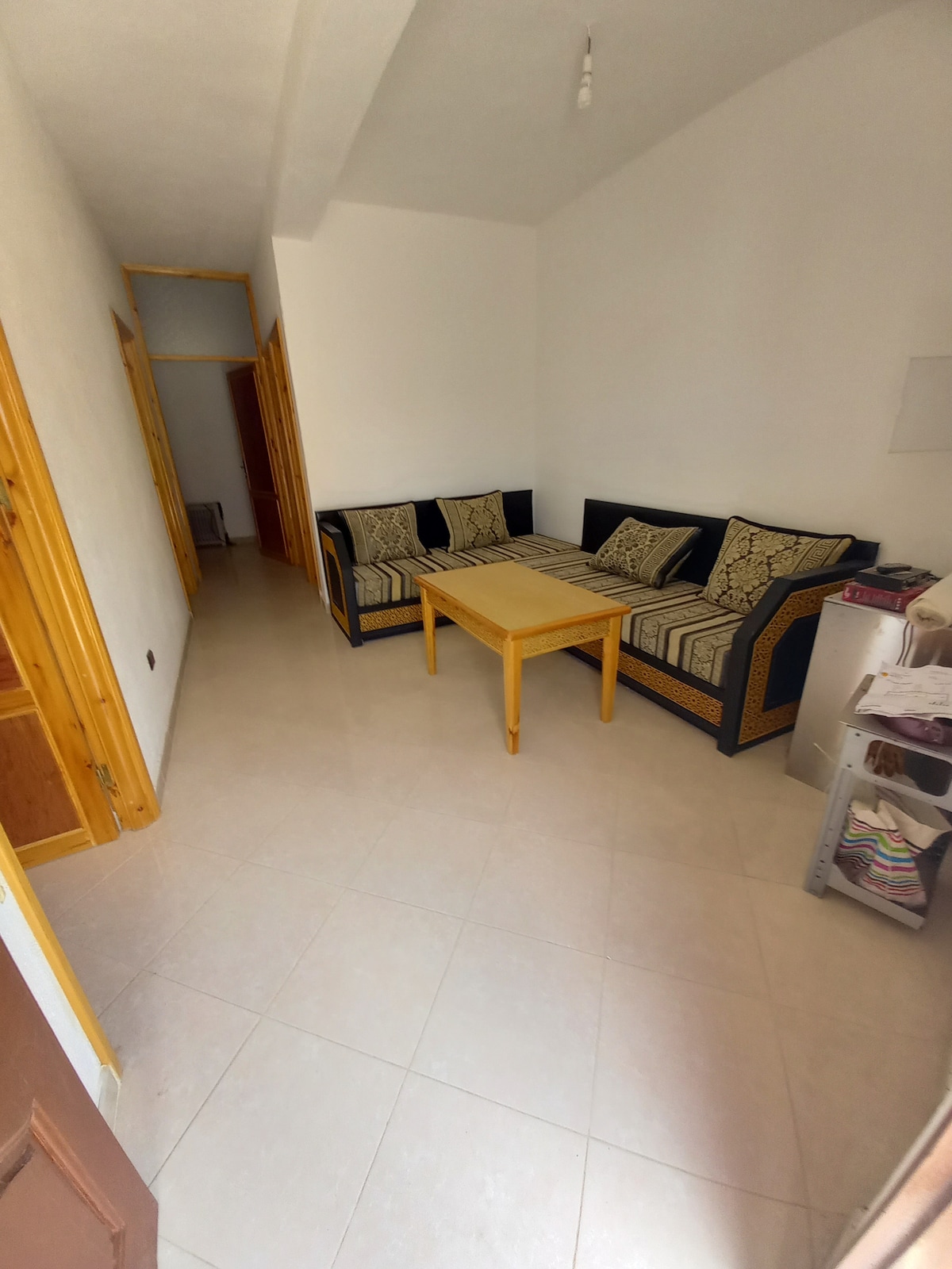 A well-lit common area is furnished with a blue sofa adorned with patterned cushions and a wooden coffee table. Light-colored tiles cover the floor, and a hallway is visible, leading to additional rooms. Natural light enters through the windows, enhancing the space.