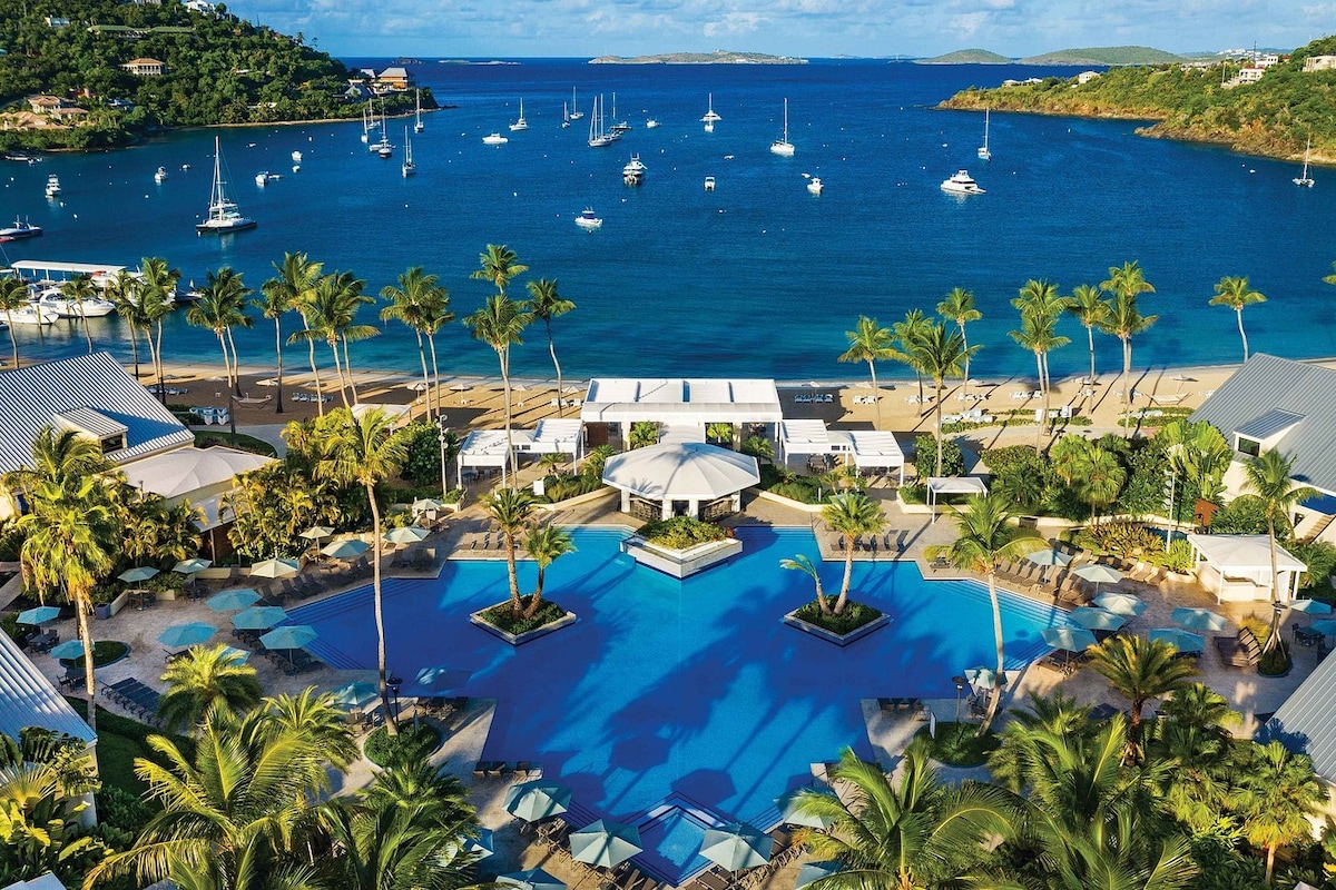 An aerial view showcases the quarter-acre pool surrounded by palm trees and sun loungers, leading to a sandy beach. Tranquil waters reflect the sky dotted with sailboats, creating a serene coastal atmosphere.
