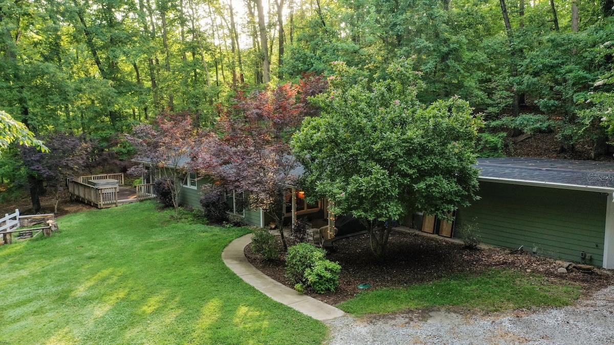 A spacious waterfront house is nestled among trees, showcasing a large, manicured yard with diverse foliage. A circular driveway leads to the home, framed by a covered porch and a deck, providing ample outdoor space for relaxation and gathering.
